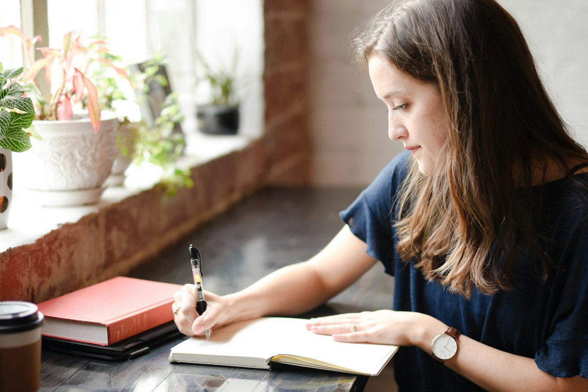 A woman writing notes in a notebook at a table beside a sunny window