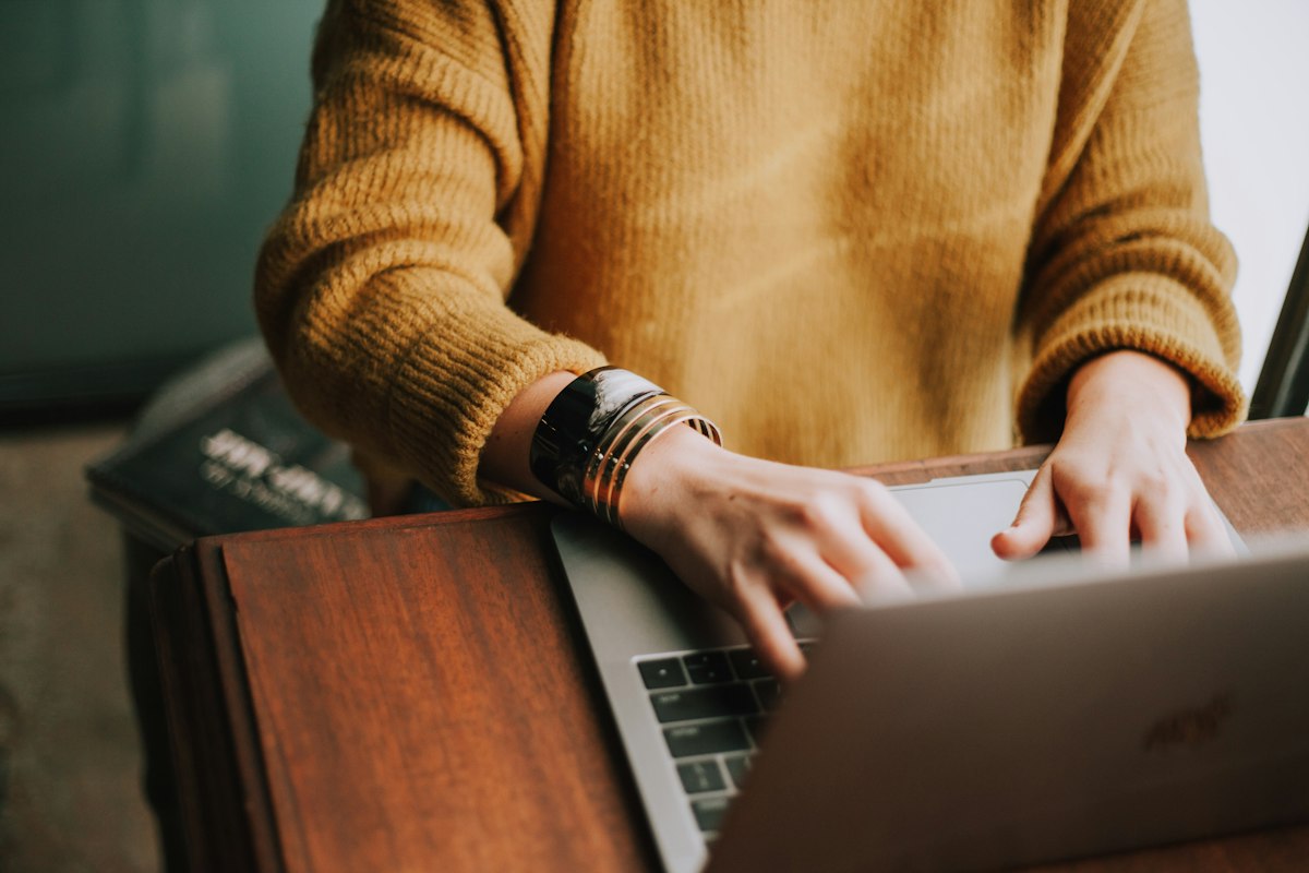 Person in a mustard yellow jumper typing on a laptop at a wooden desk while working on a side project