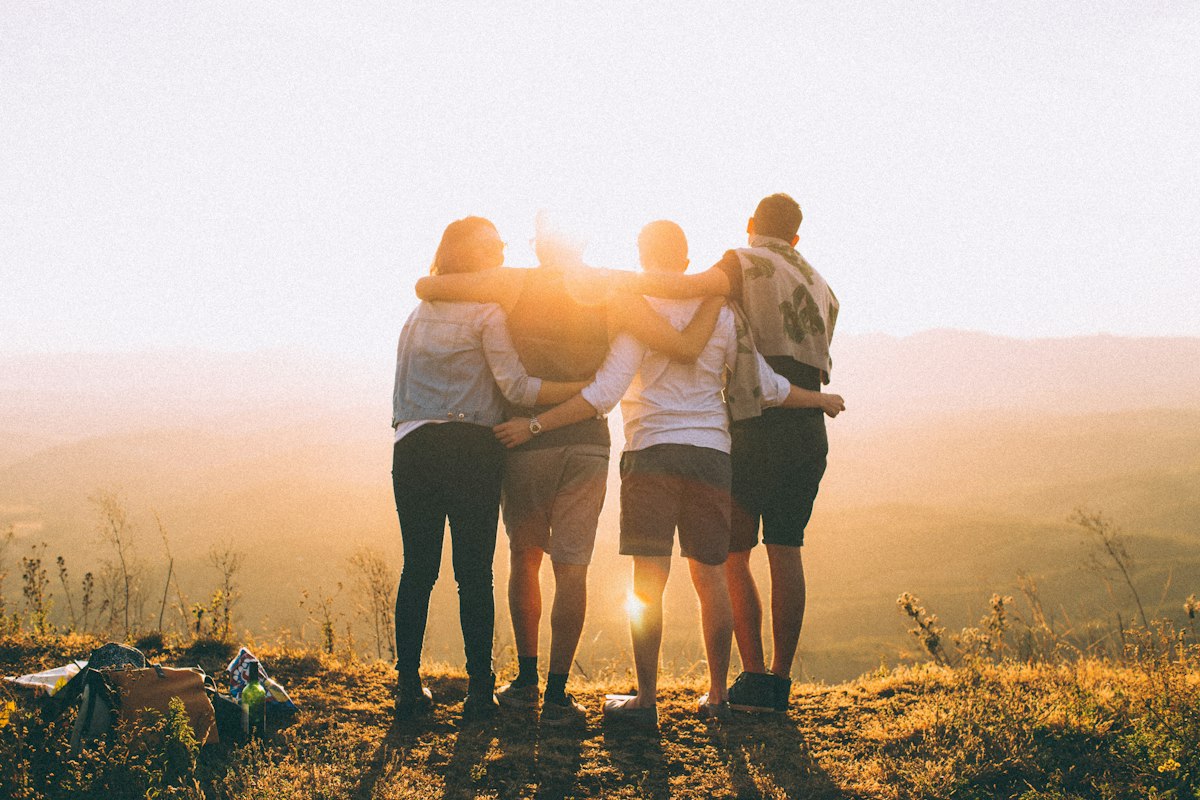 Four friends with their arms around each other watching a sunset over rolling hills