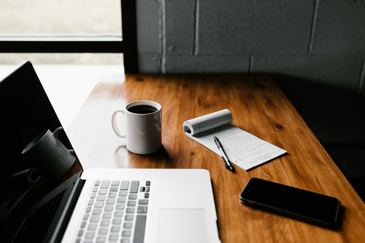 A laptop, a mug of coffee, a notepad and a smartphone arranged on a wooden desk